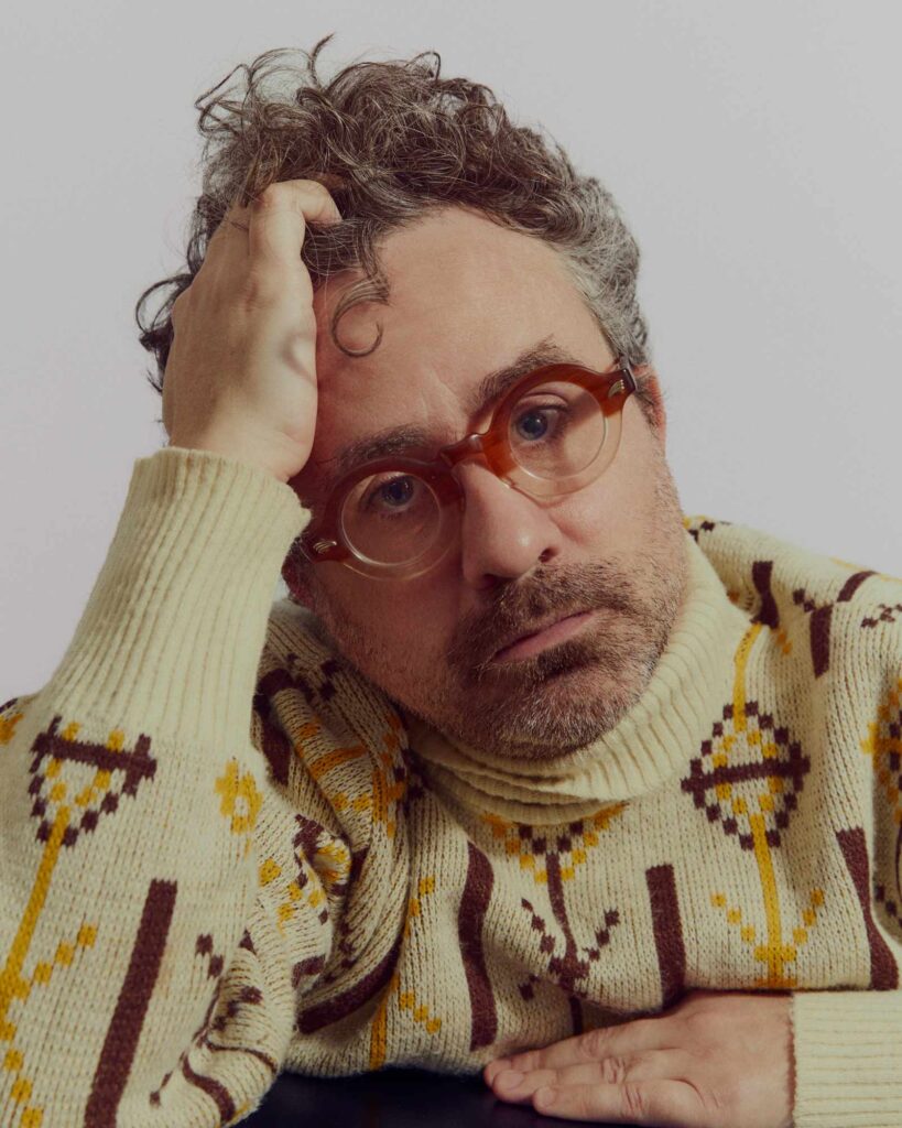 Joel Elliott wearing round glasses and a patterned sweater, looking at the camera while leaning his head on his right hand