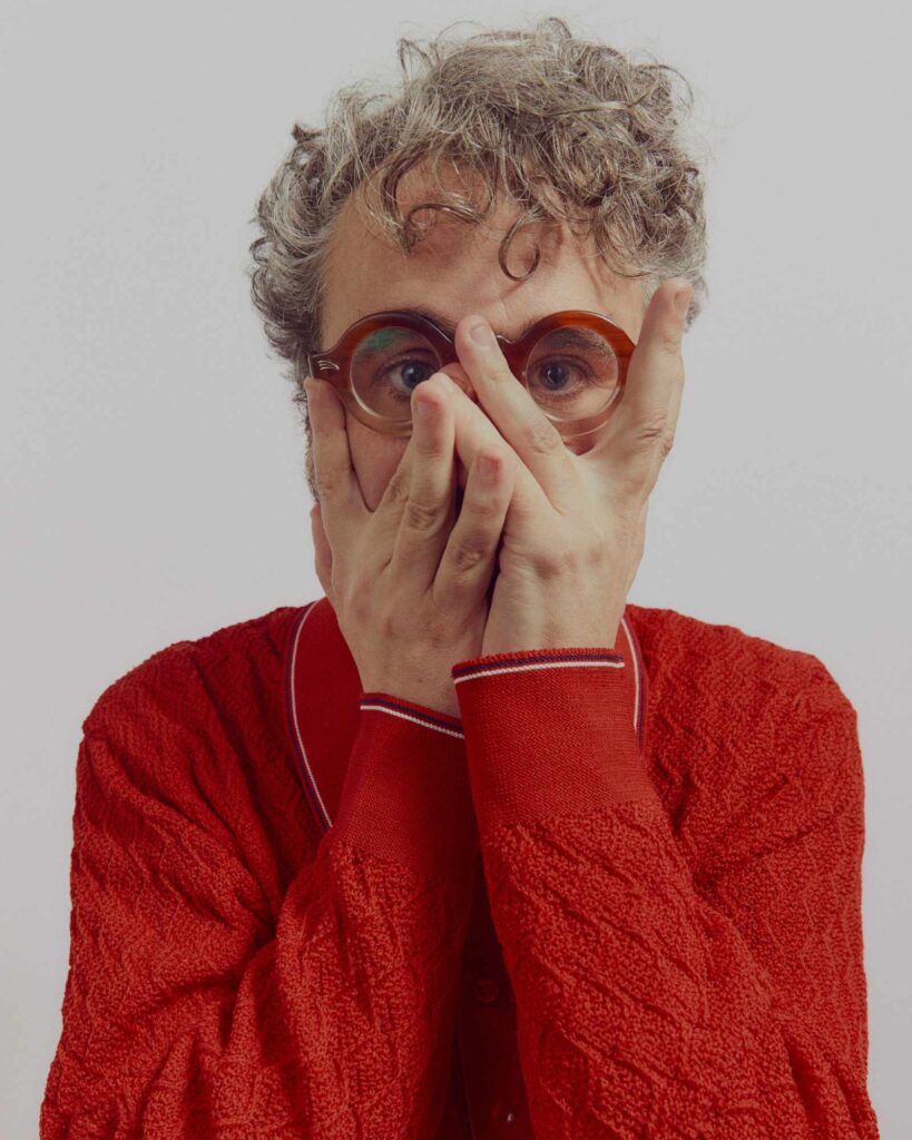 Joel Elliott, wearing round glasses and a red sweater, with his hands in front of his face